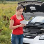 Young woman standing at broken car in meadow and reading owner manual