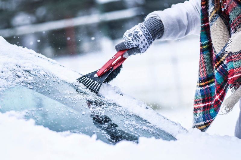 A photo of a woman scraping ice off of her car windshield in the winter.