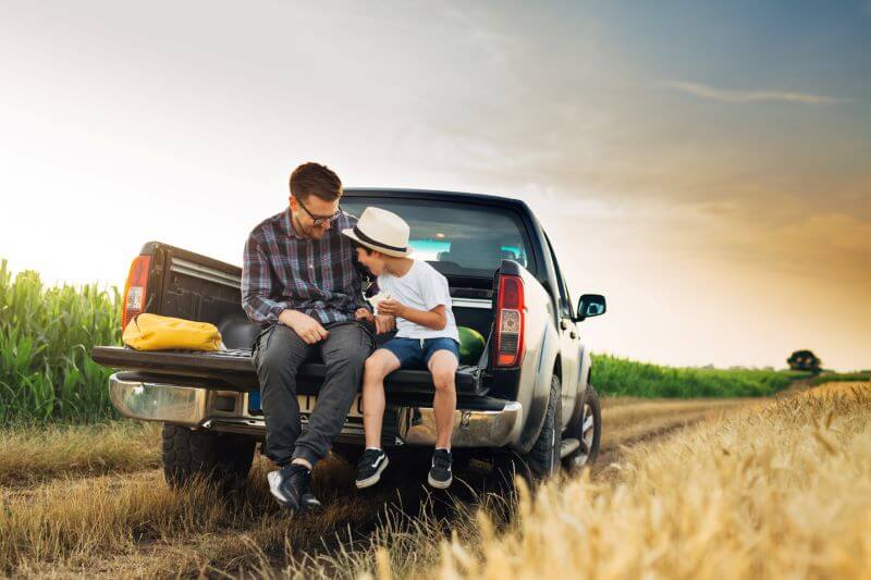 A photo of a man and his son sitting on the back of a truck in a field.